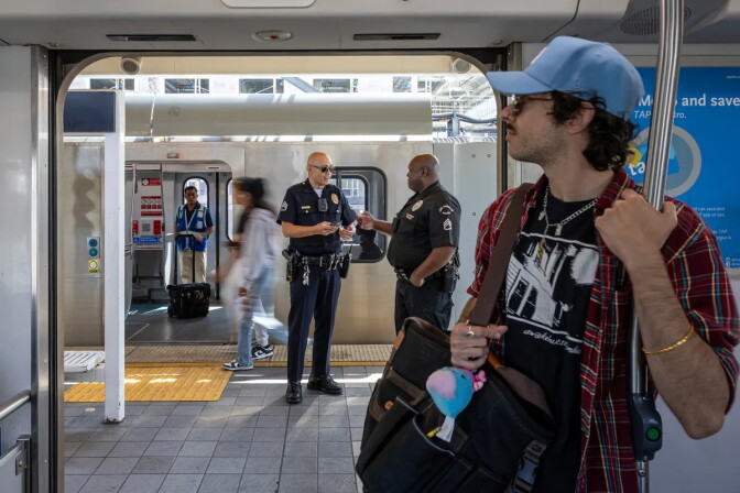 A man wearing a plaid shirt, a black tshirt underneath, and a light blue baseball cap holds onto a steel pole inside of a subway car. Through open doors, two uniformed police officers stand on the subway platform.