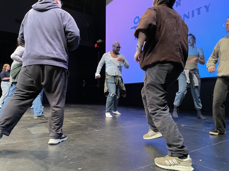People dance in a circle on a stage. The main subject of the photo is bald and wears dark-rimmed glasses. He's wearing a white undershirt with a light blue shirt over it. He's also wearing jeans.