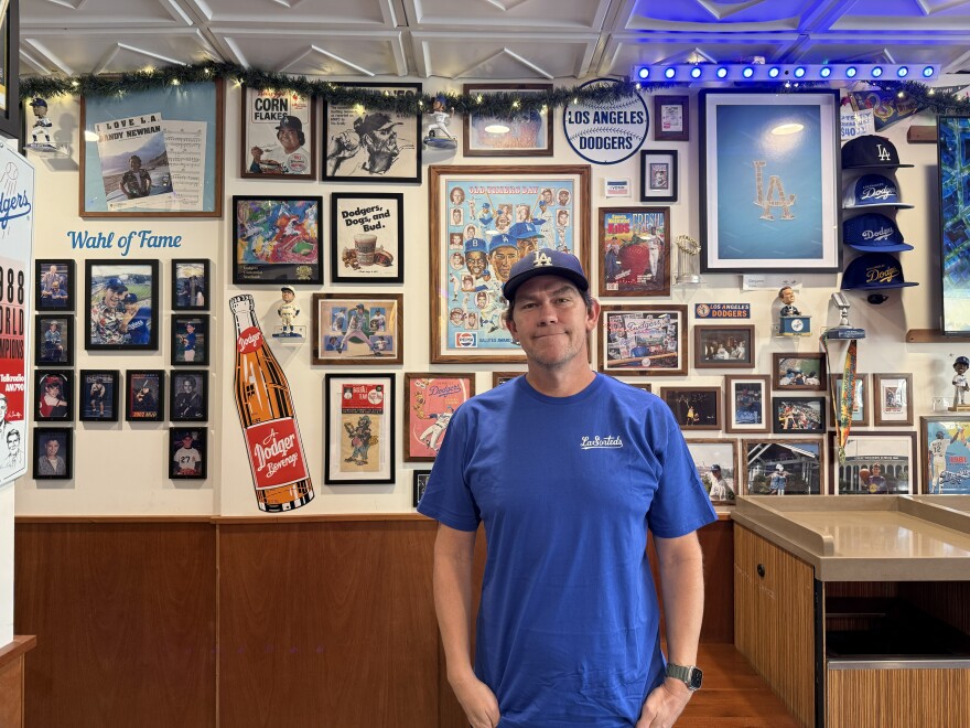 A man wearing a blue t-shirt and blue Dodgers 'LA' cap stands in front of a wall of Dodgers memorabilia that includes photos and bobble heads.