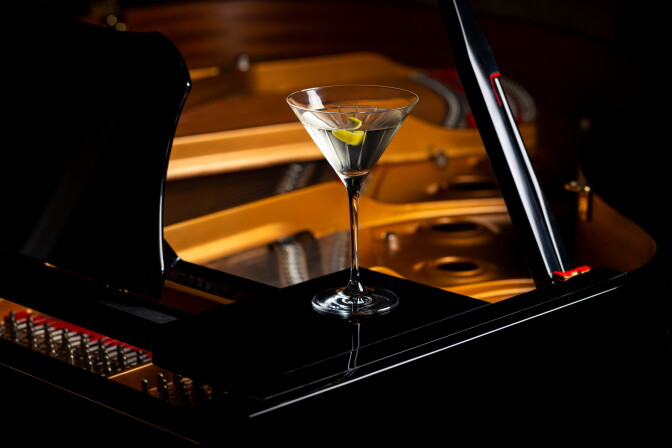 A filled martini glass sits on a glossy piano, with a warm brown glow emanating from within