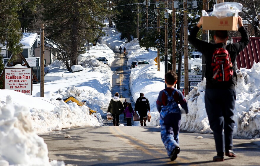 People carry boxes and bags of food down a road cleared of enough snow to let a single car drive through it. Snow is piled on either side many feet high. A sign for Roadhouse Pizza is visible on the left.
