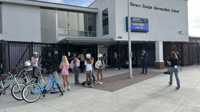 In front of a school building, a group of students stand outside. Three bicycles are parked out front. 