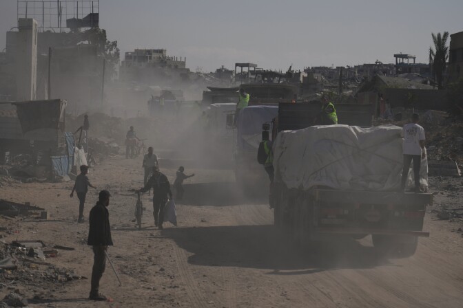 People stand along a road as trucks kick up dust.
