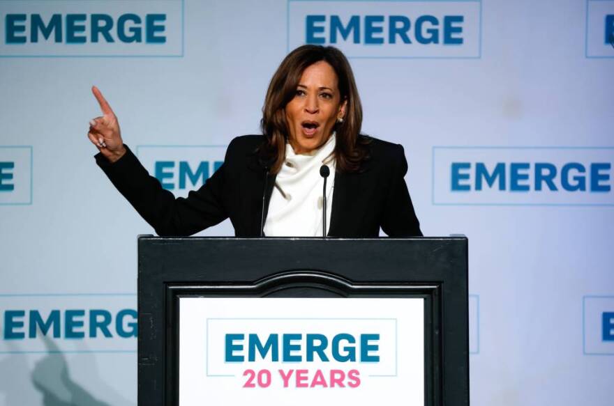 Former Vice President Kamala Harris speaks behind a black podium with signage on it that reads "Emerge 20 years" and a wall that has text that repeats reading "Emerge."