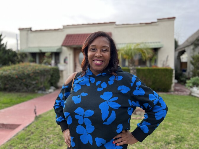 A dark-skinned Black woman in a black and blue hoodie stands in front of her house in Mid-City. 