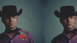 A young Jon Voight stands in front of a gray background. He wears a black cowboy hat with a black ascot tied around his neck and a light purple button down. The shirt has an embroidered red flower framed with green leaves on each of the shoulders. The image of Voight is mirrored on the other side of the frame so that he is flanking both the left and ride side of the picture. The images are from the set of "Midnight Cowboy."