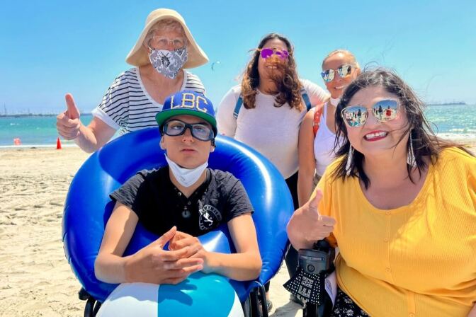 A person in a wheelchair at the beach. Four people are around him.