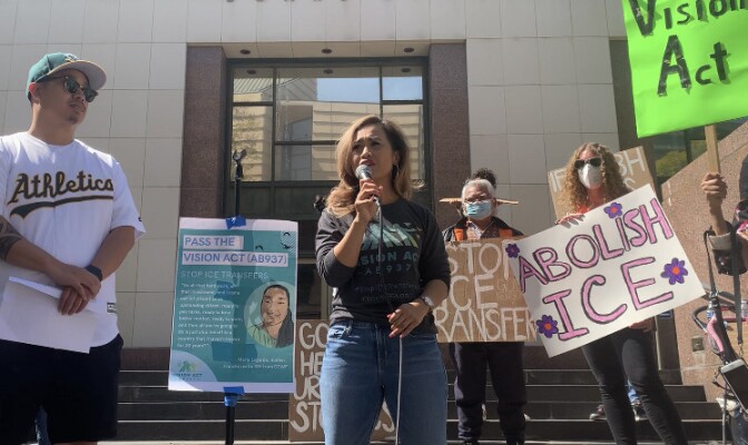 An Asian American woman stands on the steps of a building in downtown holding a mic as people around her hold signs that read "Abolish ICE" and "Stop ICE Transfers."