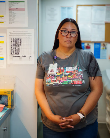 A woman stands holding her hands in front of her. She's wearing a gray graphic T-shirt and glasses, leaning against a wall.