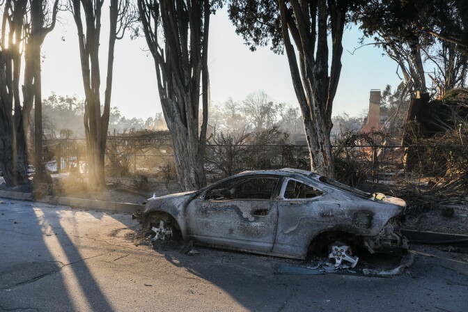 A burned out car on a sidewalk, parked in front of the burned remnants of a home.