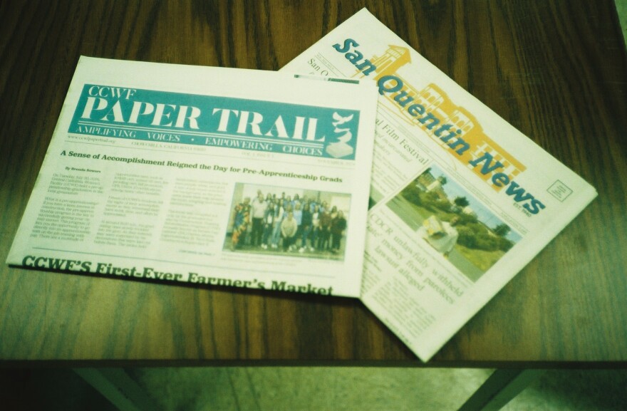 Two folded newspapers are on a dark wooden table. The first newspaper has a yellow masthead with blue lettering reading "The San Quentin News." The second newspaper is laid partially on top of the first. Its masthead is blue with white lettering and reads "The Paper Trail."