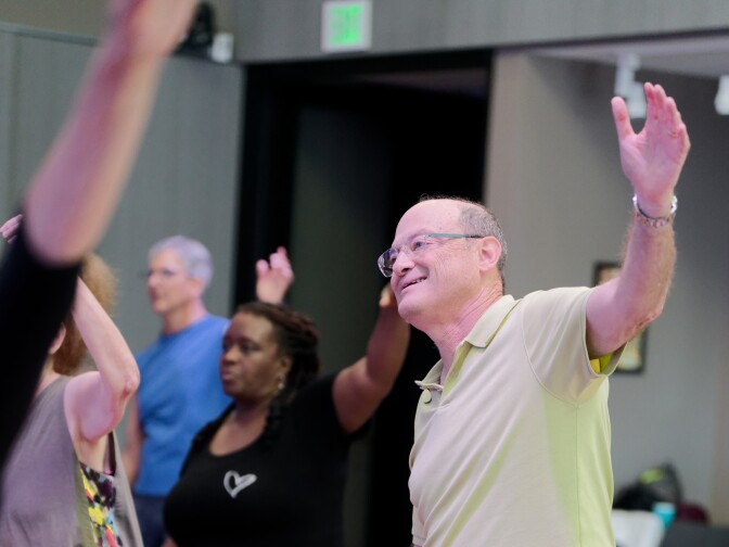 A smiling man in a light green polo shirt and blue cargo shorts participates in a group dance class. He is standing on a wooden floor with his arms raised and palms open, following the movements along with several other participants in the background.