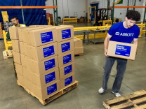 A male presenting person carries a cardboard box. A stack of about 30 boxes is next to him.