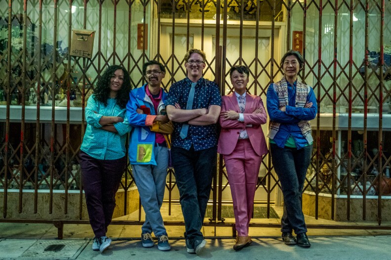 Five men and women in colorful garb stand with arms folded across their chests in a row. 