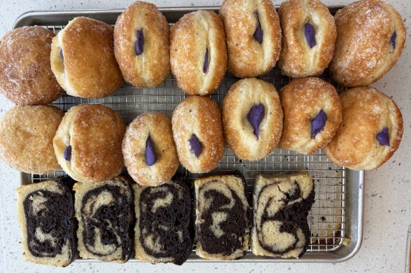 Filipino-inspired baked goods displayed on a cooling rack over a sheet tray. On the top are golden, sugar-dusted yeast-raised donuts, each filled with a generous amount of vivid ube (purple yam) cream. Below there's a sliced marble loaf cake, featuring dramatic chocolate and vanilla swirls.