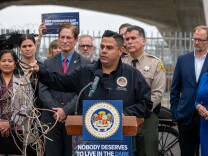 A man stands at a podium speaking into a microphone. In front of the podium, there are two signs with the text: "CALIFORNIA STATE ASSEMBLY" and "NOBODY DESERVES TO LIVE IN THE DARK." In his right hand, the man holds and assortment of tangled wires. Some are yellow, others red and white. There's a group of nine people standing behind him.