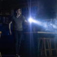 A man holds a flashlight in a dimly lit environment, surrounded by a set that appears to be a kitchen.