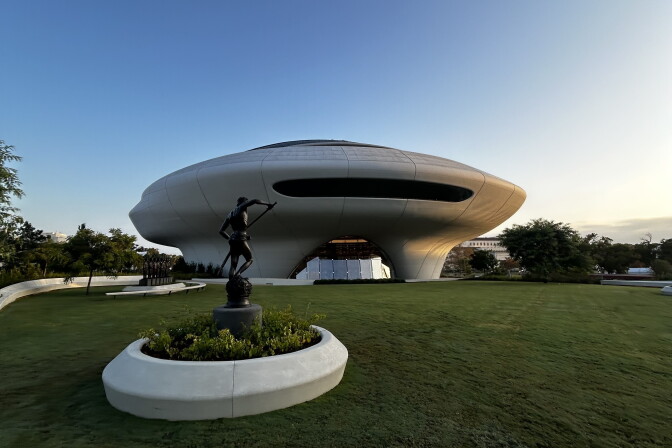 A saucer-shaped building sits on a lawn behind a bronze statue of a figure holding a pole atop a pedestal.