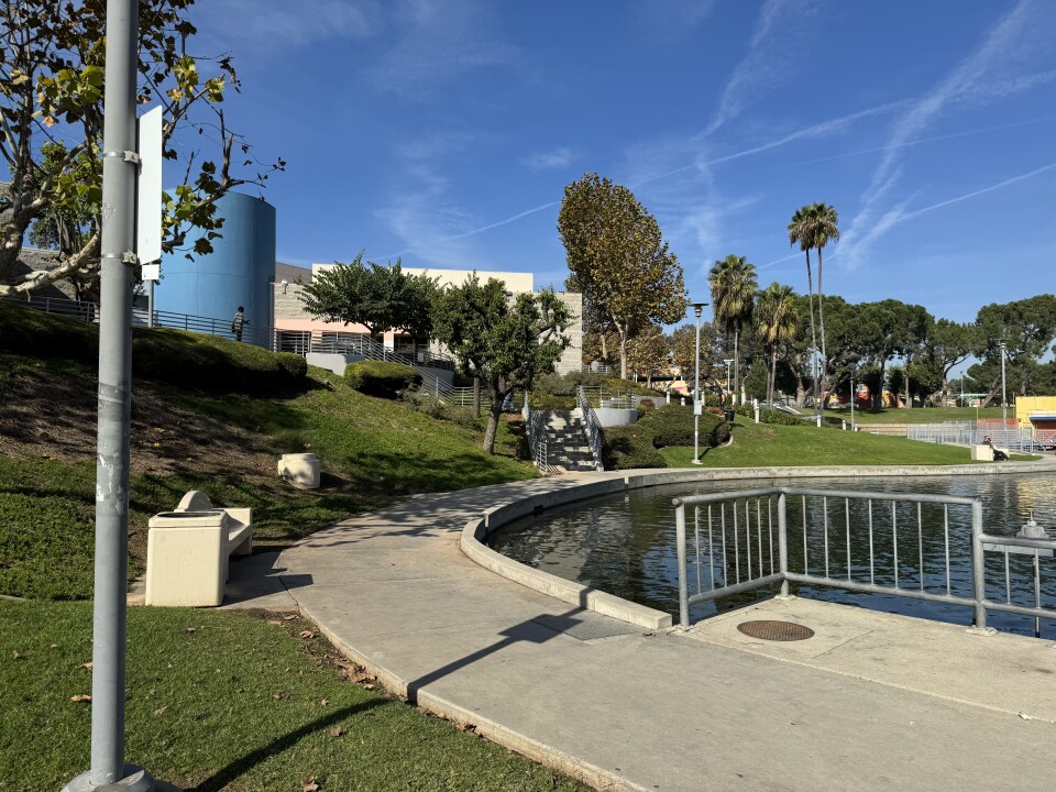  a serene outdoor setting, likely a park or landscaped public area, on a bright sunny day with clear blue skies and some wispy clouds. A building is in the background, with a big blue cylinder as part of the building.