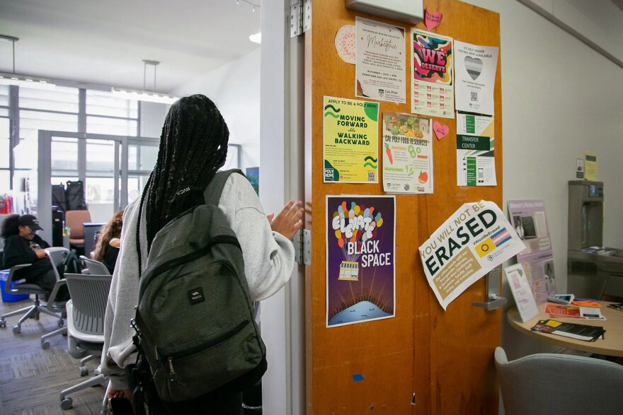 A Black student stands in an open doorway facing a classroom with their back turned. They are wearing a light grey sweatshirt and an earthy green backpack, and they have long, dark braided hair. The open door is covered in posters that say things like "Black Space" and "You will not be erased." A couple of students are sitting in office chairs in the classroom. A water bottle refill along with a round table are in the room next to the classroom, on the other side of the open door.