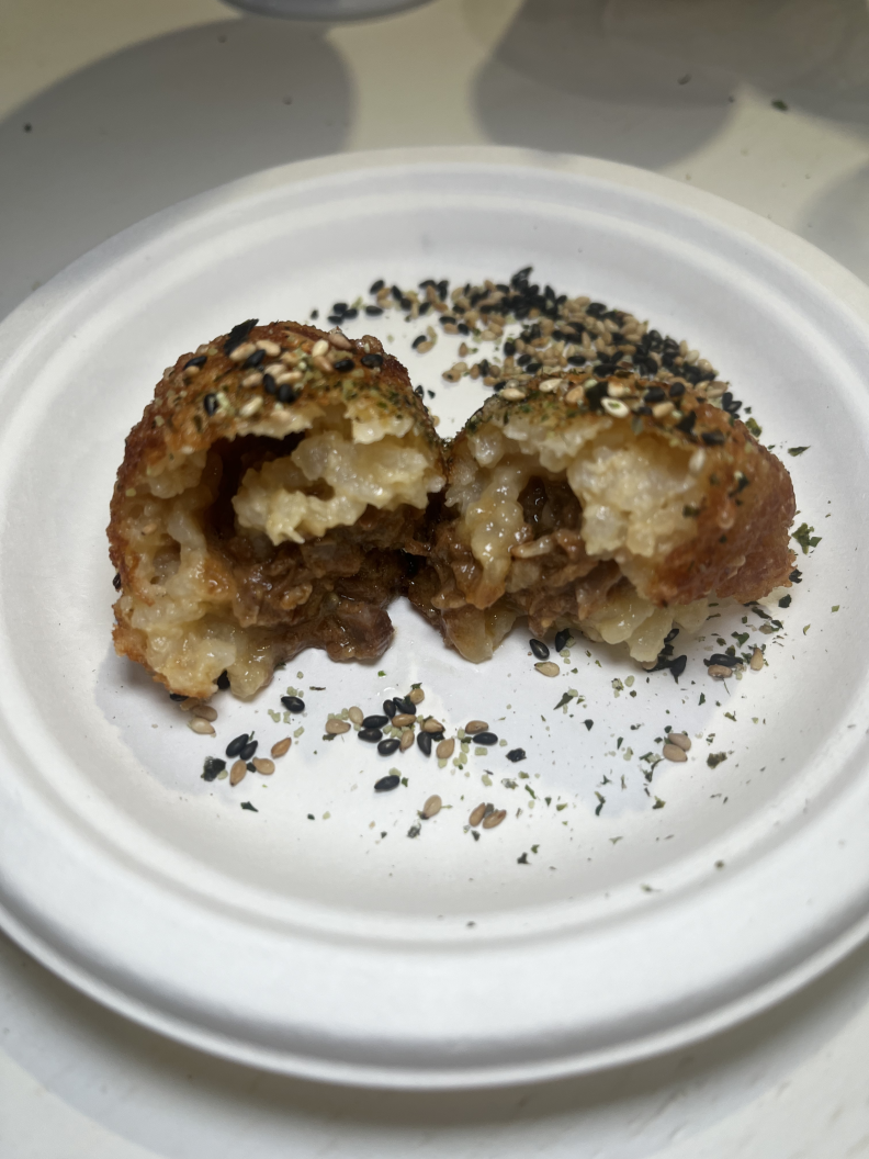 A small white paper plate holds a ball of food that is light brown on the outside and white on the inside, split down the middle. The ball contains a small amount of cooked meat and is coated with a mixture of black and white seeds.