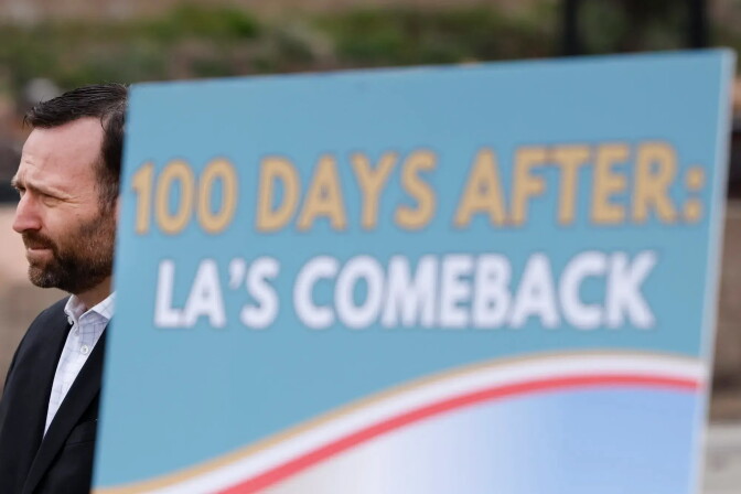 A person stands beside a sign reading '100 Days After: LA's Comeback.'


