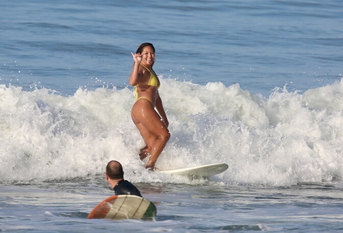 A woman with dark hair wearing a yellow bikini flashes a hang loose sign and smiles while surfing on a white board. 