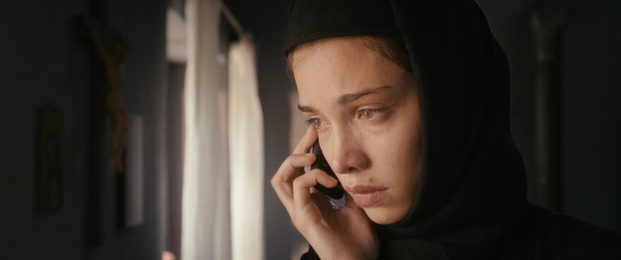 Ioana Bugarin wears a nun outfit in a shot that only shows her head. She holds a phone up to her right ear and looks off to the right, seemingly worried. In the background, a dimly lit room.