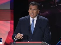 Latino man in blue suit and tie stands talking at a podium with a microphone with with Debate Night on chryron in right hand corner 