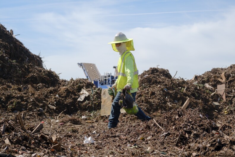 A man wearing a face covering, hard hat and neon safety vest walks through large piles of green waste collecting non-organic trash.