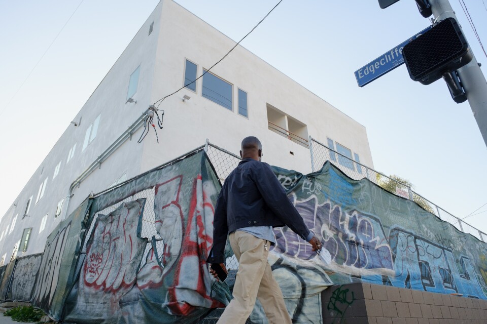A multi-story white building is surrounded by a chain-link fence. The fence is covered by a gray sheet with graffiti on it. A man in a blue coat walks by.
