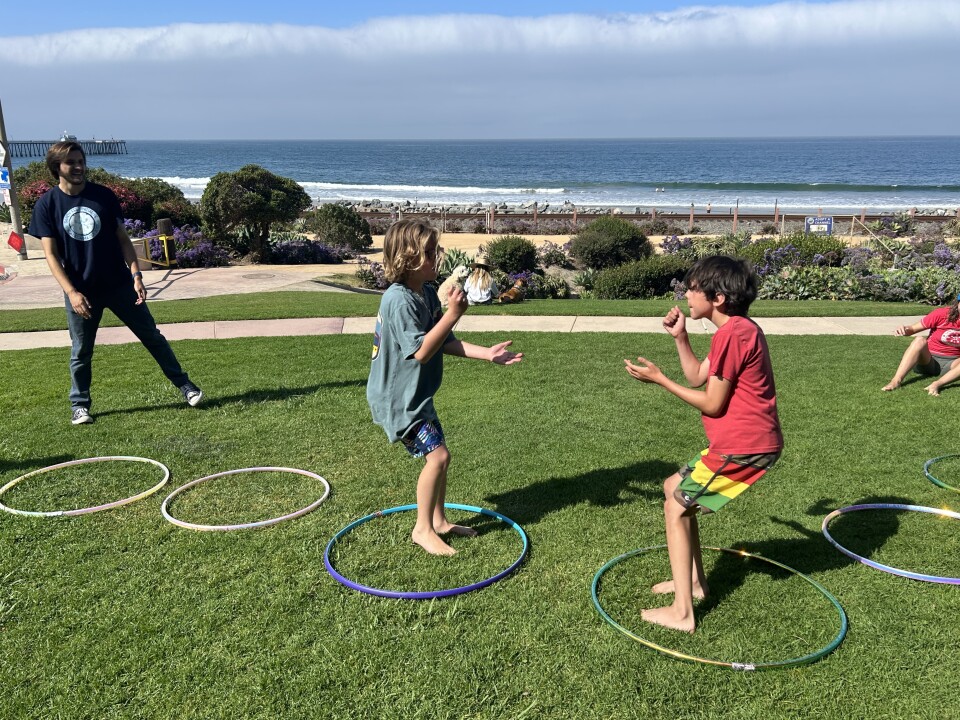 Two young male-presenting figures stand in the middle of hula hoops in a position to play rock, paper, scissors. They's on a grassy hill overlooking the ocean. 