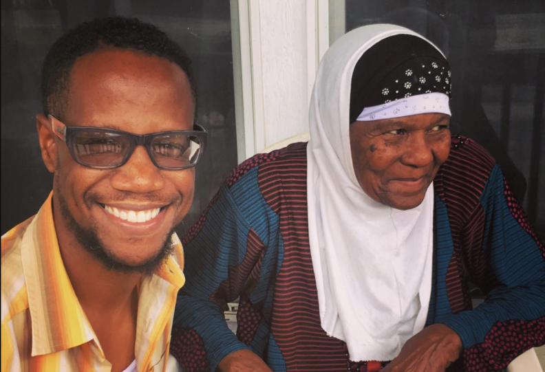 A Black man and older Black woman both smile, her hair is covered and he wear glasses and a collared shirt that's unbuttoned.
