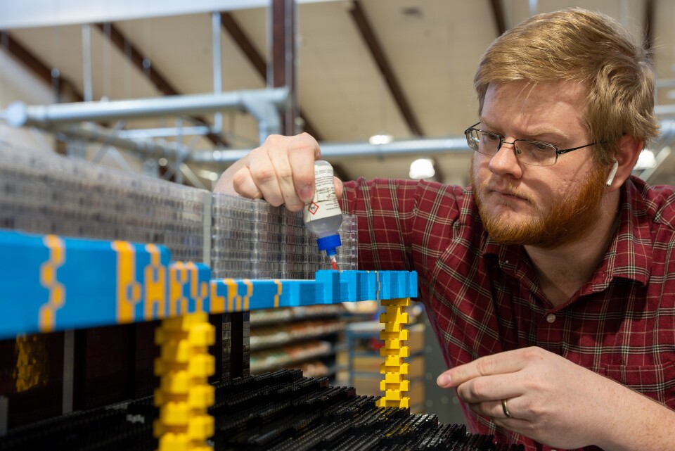 A light-skinned man with sandy hair and a beard uses glue to hold Legos together as he constructs SoFi Stadium, with large sections of blue and yellow brick columns seen in the photo. He has white Apple Airpods in his ears.