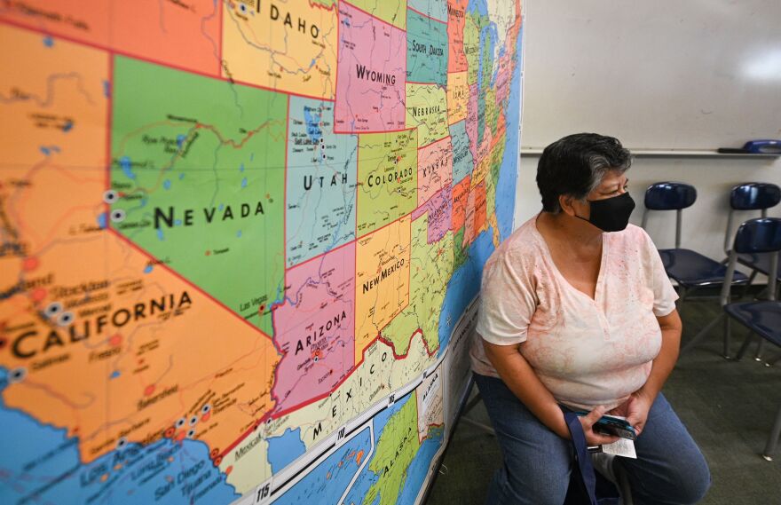 A person wears a mask while seated in front of a colorful map of the U.S.