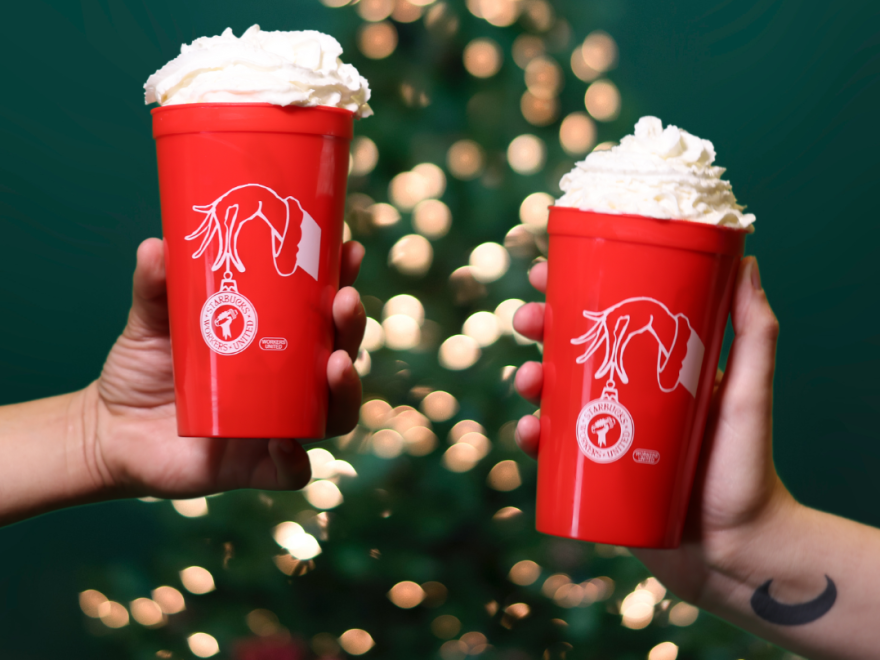 Two red plastic drinking cups are shown in close-up, each held by a person's hand. The cups appear to be full with whipped cream on top. Behind them a blurred Christmas tree can be seen.