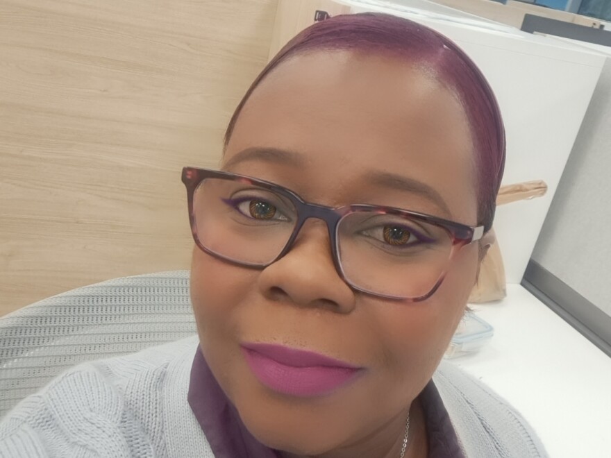 A Black woman poses for a selfie. She has dark red hair and is wearing tortoise shell glasses with purple lipstick.