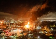 Flames rise from the center of a large refinery campus.