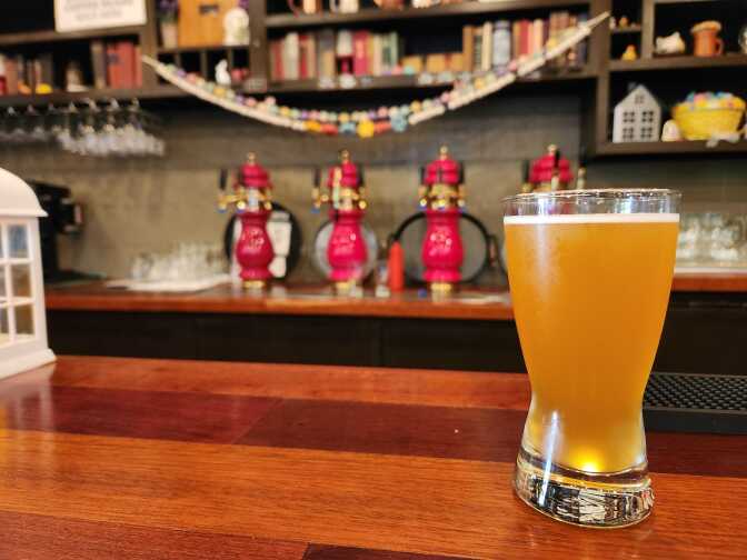 A frosty glass of golden beer, curved towards the bottom sits on a wooden bar. In the background as shiny, red beer spouts and a festive garland hangs from a shelf above filled with books. 