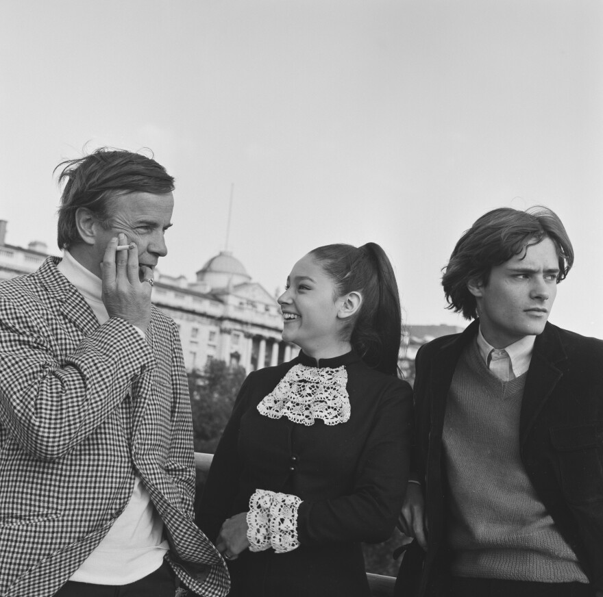 A black and white photo showing an older man on the left in a check jacket and turtle neck, a cigarette in his hand, next to a teenage girl with a pony tail smiling up at him, and a teenage boy in a jacket and sweater staring off to the right hand side.
