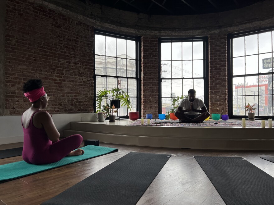 Two brown skinned people sit cross-legged in a dark studio room on yoga mats.