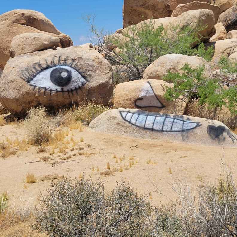 A pile of large tan rocks on a sunny, clear day. At least three of them are covered in a black and white spray painted image of a face - with a single eye, nose, and mouth each on a separate rock.