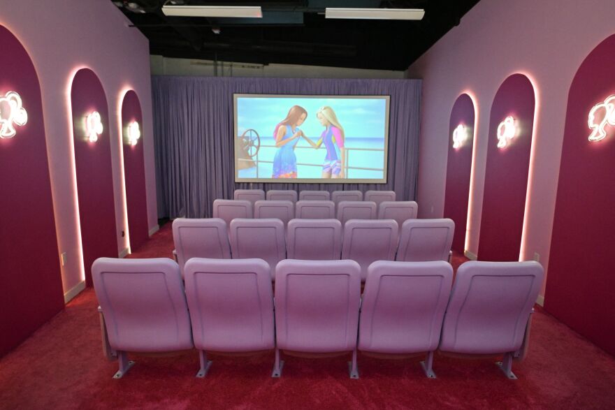 A small indoor movie theatre all in shades of pink. There are 20 viewing chairs lined up in a more pastel shade, while the walls are designed in a darker pink. A large screen is affixed to the wall, showing two Barbie dolls interacting on a boat, with the bright blue ocean waters behind them.
