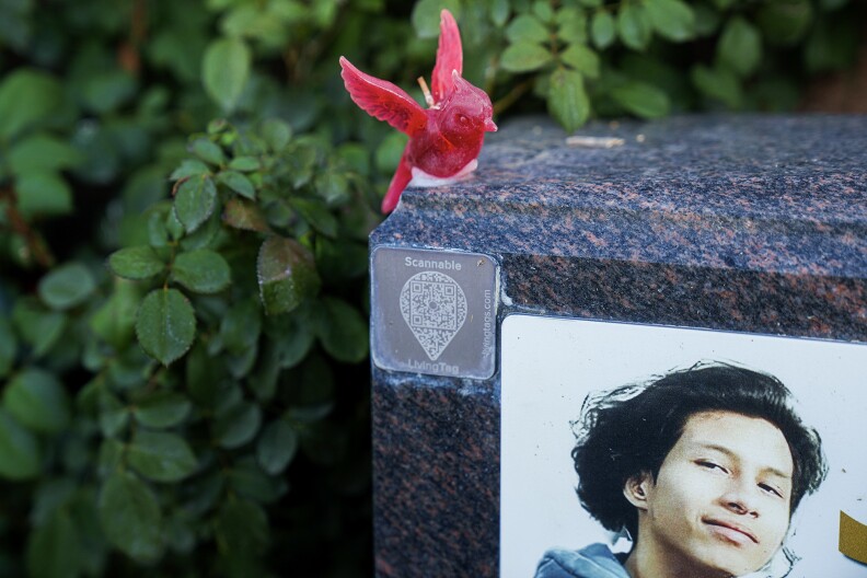 Closeup of a tombstone with a photo of a young man. A red candle in the shape of a bird is on top of the tombstone. A QR code is also attached to the front of the tombstone.