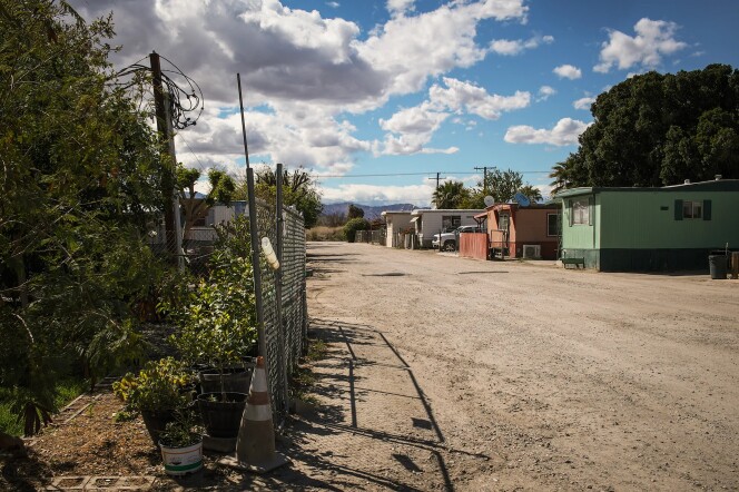 An image of a deserted dirt road. A chain link fence borders the road on the left, and several mobile homes are on the right. One is green, one is red and one is white. It is day time and the sky is white with clouds. 
