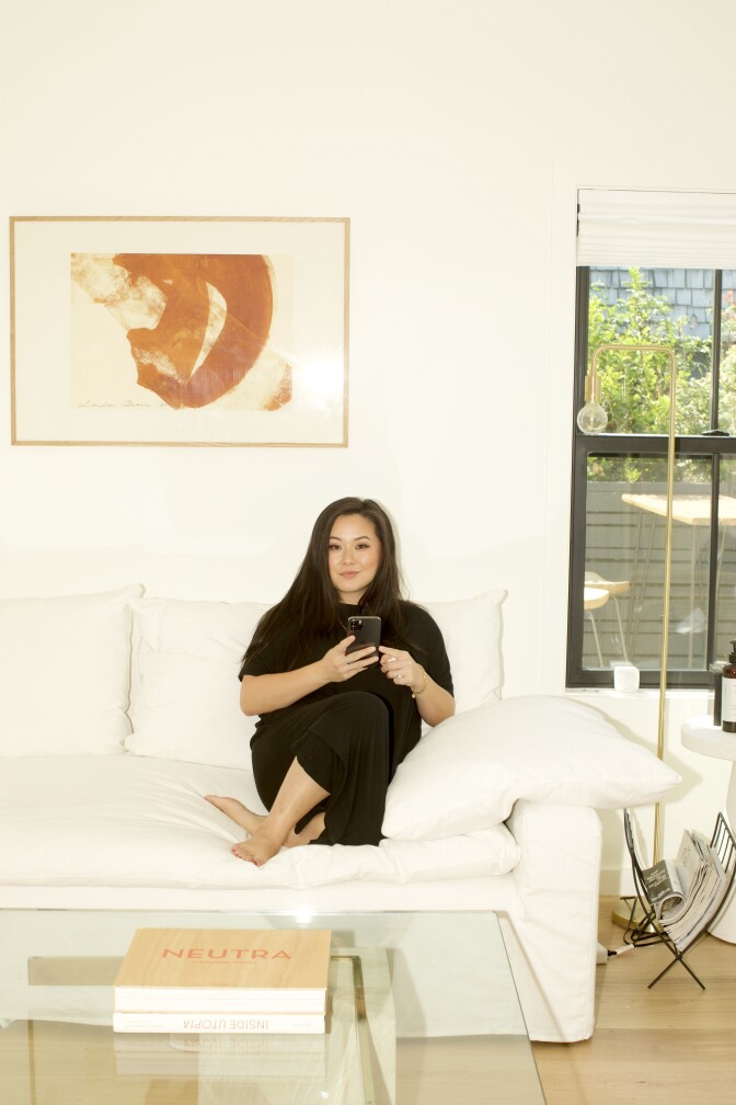 Jing Gao, a young Asian woman with black hair, is sitting on a white couch in her living room, dressed entirely in black attire. She is barefoot with her legs slightly crossed, holding a black iPhone. Above her, there is an abstract painting hanging on the wall, and next to her is an open window that lets in the sunshine. In front of her, there is a lucite coffee table with two large coffee table art books stacked on top of each other.