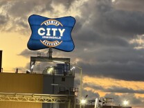 A big blue sign that say "City Brewing & Beverage Irwindale" is seen at sunset. Lights illuminate the sign. Large clouds can be seen behind it. Palm trees and power lines are seen in the right hand corner.