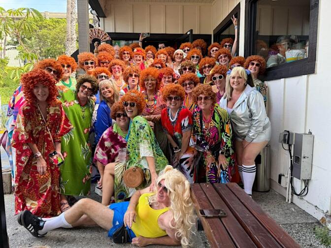 A large group in colorful outfits and red, curly wigs posing outdoors. One person lies in front wearing yellow top and blue shorts.