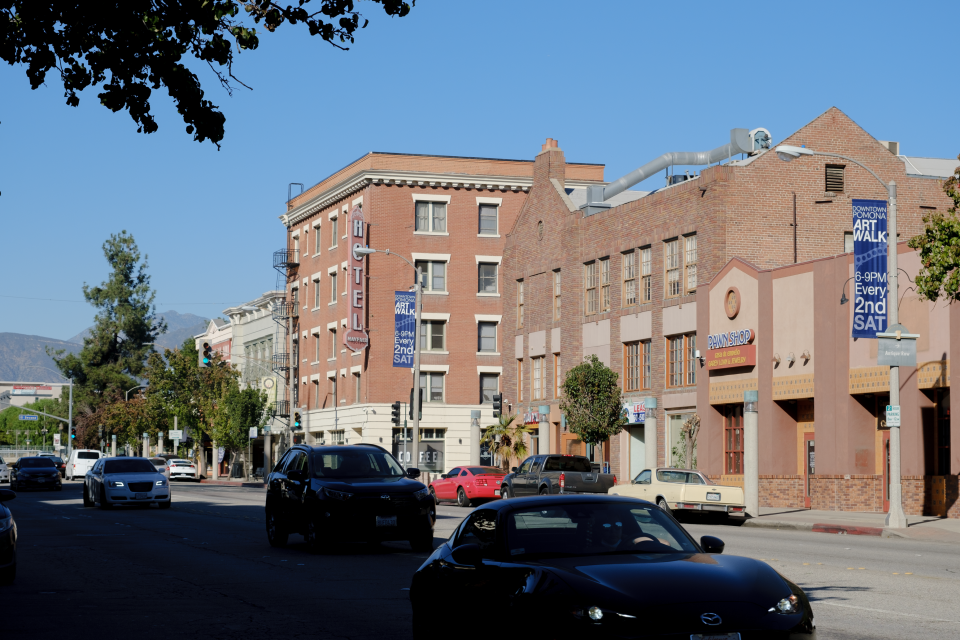 There is a row of brick buildings in what looks like a small-town downtown area. A few cars and some foliage are silhouetted in the foreground.