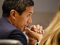A closeup photo of a man in a suit jacket looking ahead while holding his thumb up to his chin while clasping his hands around a pen.
